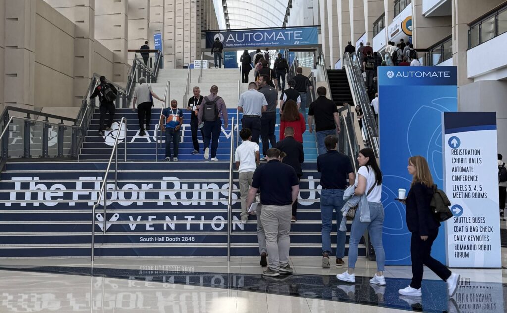 Crowd of conference attendees walking up a wide blue-carpeted staircase under an AUTOMATE banner in a large atrium.