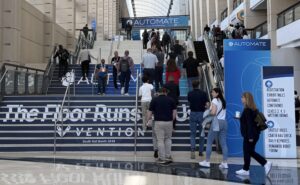 Crowd of conference attendees walking up a wide blue-carpeted staircase under an AUTOMATE banner in a large atrium.